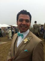 A man wears a tan suit with a flower pinned to his lapel and smiles at the camera, at what appears to be a formal attire outdoor event. Behind him, people dressed in nice clothes walk on a grassy lawn between tents and awnings.