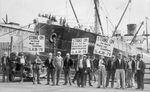 A black and white image of men holding signs with messages about a strike who stand on a dock, with a large boat or ship visible behind them.