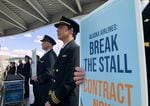 People in pilots' uniforms stand side by side. One person holds a sign that reads ""Alaska Airlines: Break the Stall Contract Now"