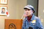 An older person wearing a denim jacket and a cap speaks while standing in front of a podium that has a Japanese flag on one corner and a and U.S. flag on another.