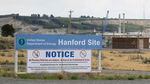 A sign reads "U.S. Department of Energy Hanford Site" and includes warnings that people and vehicles may be searched for prohibited items. In the background, a building and a crane can be seen.