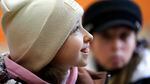 A 10-year-old girl wearing a white beanie is photographed in profile. She is smiling, in focus. Behind her, another person in a black hat is out of focus.