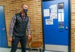 principal wearing a black vest and plaid shirt, walking past a blue classroom door that is marked with a notice stating isolation area