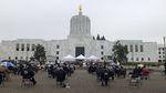 People sit outside in chairs on a gray day facing the Oregon State Capitol building for an event.
