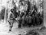 A  U.S. soldier of the 12th Armored Division stands guard over a group of Nazi prisoners captured in the surrounding German forest. April 1945.