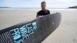 A man sits for a photograph with his black surfboard on a sandy beach. The board has many names on it in white letters and the words "One Last Wave Project" in larger blue letters.