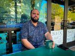 A black man sits on a patio table with a green mug of coffee.  A glass storefront is behind him, where there is a sign that reads: "BLACK LIVES MATTER."