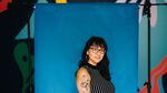 A portrait of Portland writer Emilly Prado. She's pictured from the waist up, standing in front of a blue photography background set. Behind the hanging background, a colorfully painted wall can be seen. Prado is Chicana, is wearing her black hair down with bangs, glasses, hoop earrings and a black and white striped tank top. Her right arm, facing the camera, is decorated with old-school style tattoos. She gives a slight smile at the camera.