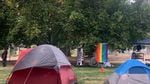 Colorful tents are pitched in a grassy city park. In the background, a flag reads "Black Lives Matter," next to a rainbow pride flag.