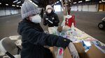 A woman in a hoodie, gloves and a winter hat reaches into a box of vaccine supplies while sitting at a table in a large, empty building. Another worker stands at the foot of the table while preparing doses.