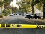 Police tape blocks a tree-lined street where cars are parked. A police car is visible in the distance.