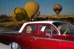 A woman and a man ride in a vintage car as hot air balloons float behind them in the region of Cappadocia, Turkey.