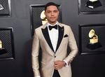 Trevor Noah poses for a photo in front of a black wall, filled with framed photos of a golden grampahone, while wearing silvery, striped tuxedo with a bowtie.