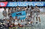 Members of the Gonzaga basketball team pose for a photo, huddled together around a banner reading "WCC 2021 West Coast Conference Champions" as confetti falls from the sky. An LED sign in the background reads "Bulldogs Win" with the Gonzaga mascot logo.