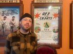 Eric Isaacson stands next to the hand-drawn event poster for the Off the Charts screening and song contest in the lobby of the Hollywood Theatre. He's wearing a yellow and black plaid flannel and a black beanie.