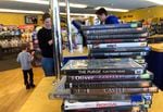 A stack of DVDs sits on a counter, while an adult and a child in the background check out at a register and a clerk stares at a computer.