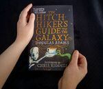 Northwest student Linn Jaster McCormick holds a book that's meaningful to them: The Hitchhiker's Guide to the Galaxy. A close up view from above of two hands holding a copy of the book against a black background. The title is in gold lettering and underneath is an illustration of a robot and a green tentacle arm holding, on either side, a teacup with gold lines swirling out of it.