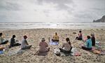 Eight women sit in a semi-circle on the beach while a ninth woman leads them in a yoga session.