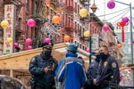 Three masked NYPD officers, all in uniform, talk with an older man dressed in a winter coat and a bucket style hat. They're standing on a street in New York City's Chinatown, decorated with lanterns hanging on wires draped above the street.