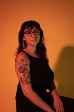A portrait of Portland writer Emilly Prado. She's shown seated against an orange background, her shadow on the wall. She is Chicana, has black hair with bangs, and wears glasses, earrings, and a black tank top. Her right arm, facing the camera, is decorated with old-school style tattoos. She gives a serious but peaceful look into the camera.