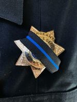 A close up image of a police badge, partially covered in a black ribbon with a blue stripe down the middle.