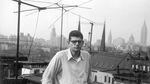 Beat poet Allen Ginsberg, photographed in black-and-white on a rooftop by William S. Burroughs in 1953. The young Ginsberg wears thick framed eyeglasses, a white button-up shirt with the sleeves rolled up, slacks and a serious expression. Behind him in view are rooftops and a distant cityscape.