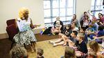 A drag performer in a blonde wig reads the children's book "Rainbow Fish" to a group of kids and parents at a library.