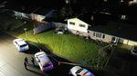 A photo taken from above shows police tape cordoning off a yard and house. Inside the tape, tire tracks show the path a car took before it crashed into the fence of the house. Outside the tape, three police vehicles are parked, and three officers stand in the street and talk to one another.