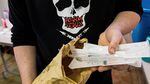A person wearing a shirt with an image of a skull and the words "Harm Redux" printed on it holds sealed and packaged syringes in one hand.