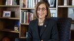 Kate Brown wears a suit and sits in an armchair in front of a wooden bookcase.