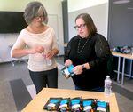 Mary Stevens-Krogh, the district coordinator of substance use supports at Portland Public Schools, stands to the left of a wooden table, holding a water bottle. Clair Raujol, addictions prevention coordinator at Multnomah County, opens a Deterra drug-deactivating bag, with more bags displayed on the table.
