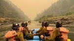 Four firefighters holding paddles in a boat, gazing into an orange-hued, smoky sky.