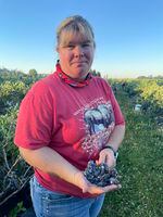 A woman stands outside in a field and holds a cluster of blueberries in the palm of her right hand.