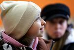 A 10-year-old girl wearing a white beanie is photographed in profile.  She is smiling, in focus.  Behind her, another person in a black hat is out of focus.