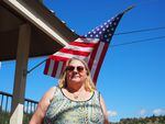 A woman in a sleeveless top stands in front of an American flag that hangs against a bright blue sky.