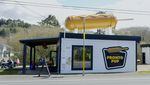 A roadside building painted white with black trim bears a sign saying "The Original Pronto Puppy" with a giant fiberglass corn dog on its roof.