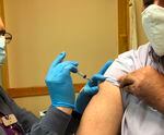 A person wearing a mask and gloves injects vaccine into the arm of a person with a rolled up sleeve who is also wearing a mask.