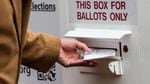 A hand puts a ballot envelope into a slot in a box with text that reads "This box for ballots only."