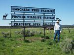 A man in a cowboy hat stands in front of a tall sign that reads "Ranchers feed families, provide wildlife habitat, preserve our open spaces."