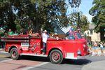 A vintage 1950s fire engine is driving down the street in a parade. Standing up front in the fire engine is Pastor Luis Molina, a man wearing a white cowboy hat, a white button up shirt, jeans and cowboy boots. He's smiling and waving toward the camera.