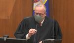 A person in a judge's gown speaks into a microphone while wearing a mask.