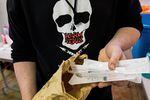 A person wearing a shirt with an image of a skull and the words "Harm Redux" printed on it holds sealed and packaged syringes in one hand.