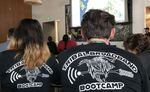 Two people sit in a room with their backs to the camera. The backs of their black T-shirts read "Tribal Broadband Bootcamp" in white letters.
