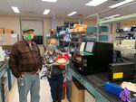 Two people stand in between lab benches on which is stacked lab equipment, gloves and paperwork. Both are wearing masks