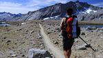A hiker carrying a backpack looks towards a dusty trail, mountains and blue sky.