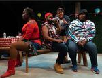 Four BIPOC actors on a stage sit together around a picnic table while wearing informal clothing.