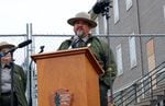 A man with a salt-and-pepper goatee and wearing a ranger's outfit speaks at a podium in front of a chain-link fence, where there is a construction site.