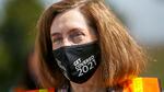 Governor Kate Brown wears an orange traffic safety vest and a mask that has the words "Get covered 2021" printed on it.