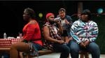 Four BIPOC actors on a stage sit together around a picnic table while wearing informal clothing.