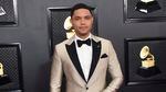 Trevor Noah poses for a photo in front of a black wall, filled with framed photos of a golden grampahone, while wearing silvery, striped tuxedo with a bowtie.